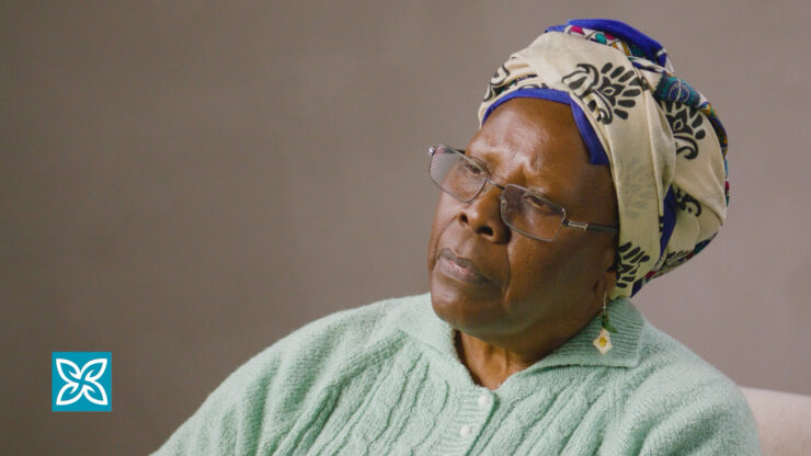 Elderly woman with glasses and a patterned headscarf sits in a chair, wearing a light green knit sweater.