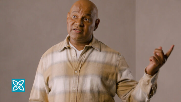 A man is speaking, with his index finger pointing up. He is wearing a plaid shirt against a plain background.