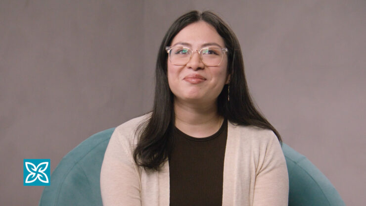 Smiling woman with dark hair wearing a brown top with a beige cardigan against a neutral background.