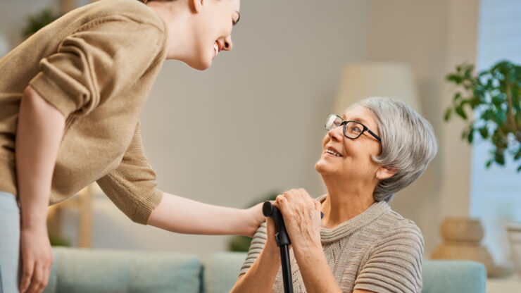 A smiling elderly woman with glasses, holding a cane, sits on a teal sofa and looks up at a young woman leaning toward her.