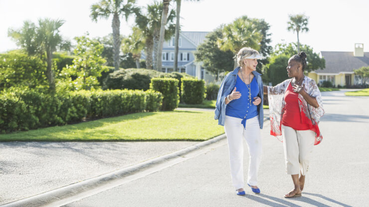 Two women walk and chat on a sunny street lined with greenery and palm trees. One wears a blue outfit, the other red.