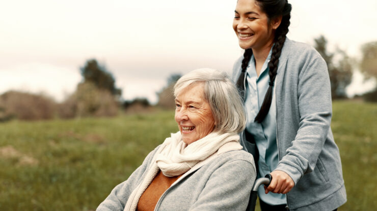 An elderly woman in a wheelchair happily smiles while a young caregiver with braided hair pushes her outdoors in a grassy field.