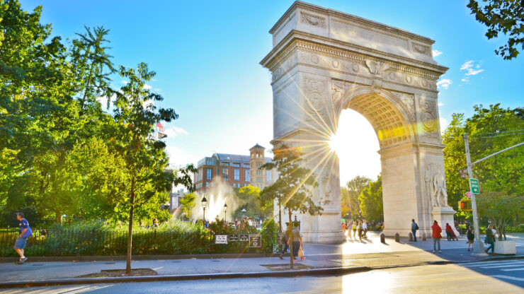Washington Square Arch in Lower Manhattan with sunlight streaming through, surrounded by trees and bustling parkgoers.