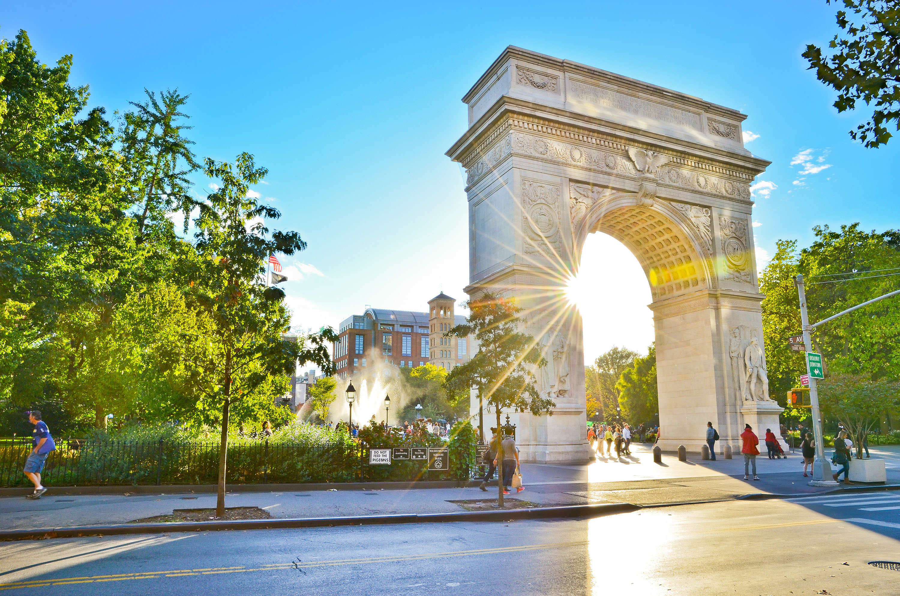 Washington Square Arch in Lower Manhattan with sunlight streaming through, surrounded by trees and bustling parkgoers.