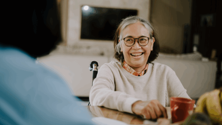 Elderly woman with glasses and a joyful expression sits at a table, holding a red mug. She is conversing with a person in a blue shirt.