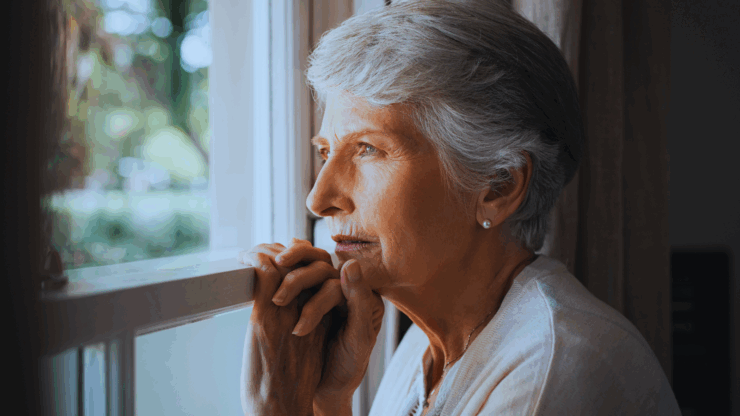 Elderly woman gazes thoughtfully out a window, her hands gently clasped. Soft natural light highlights her serene expression and gray hair.