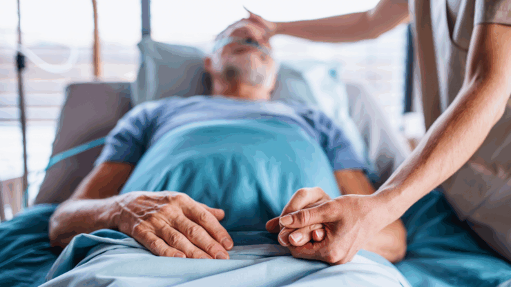 An elderly man in a hospital bed with an oxygen tube, holding hands with another person.
