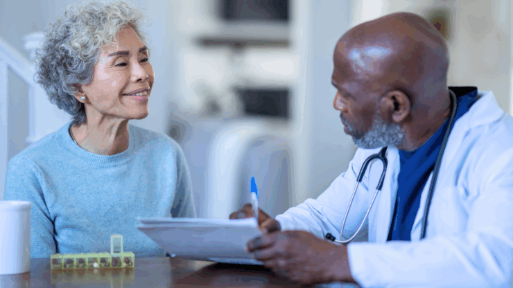 An elderly woman smiles warmly at a doctor in a white coat, holding a clipboard. A pill organizer and mug are on the table. The scene conveys trust.