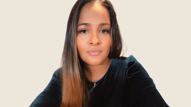 Woman with long, straight hair and calm expression, wearing a dark top and cross necklace, against a plain background.