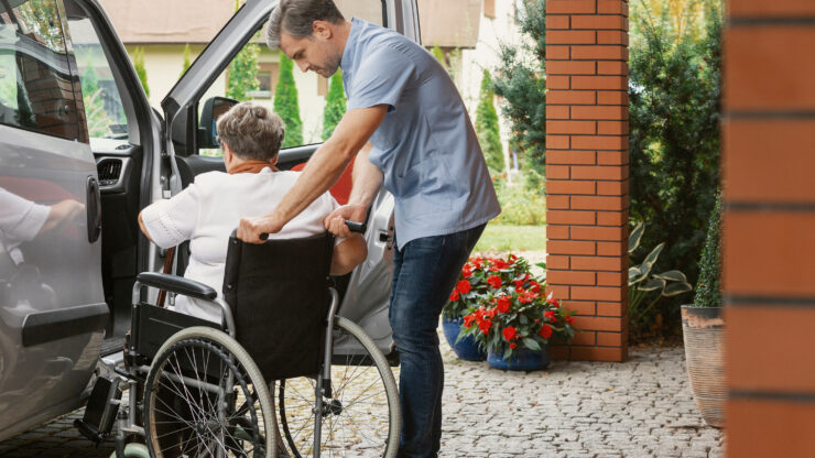 Caregiver assisting an older adult in a wheelchair into a vehicle outside a residential home.