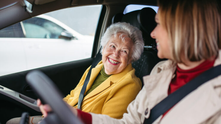 Two women share a joyful moment in a car. The older woman, wearing a yellow coat, smiles warmly at the younger woman, creating a lighthearted atmosphere.