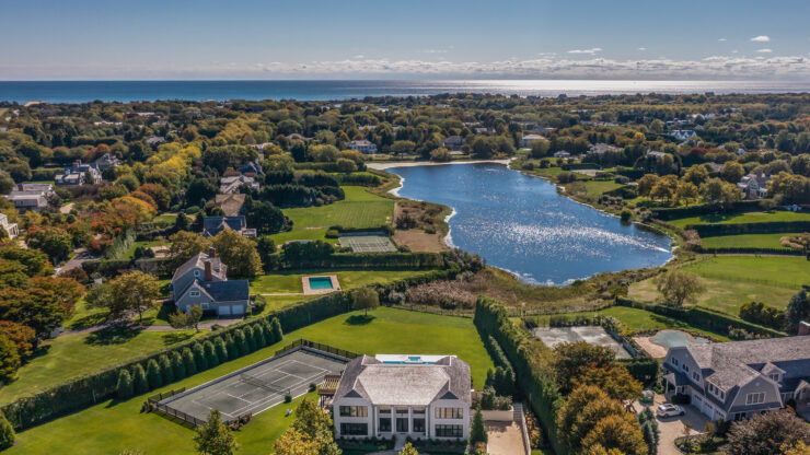 Aerial view of a coastal estate with a large house, tennis court, and pool surrounded by lush greenery. A shimmering lake and ocean can be seen in the distance.