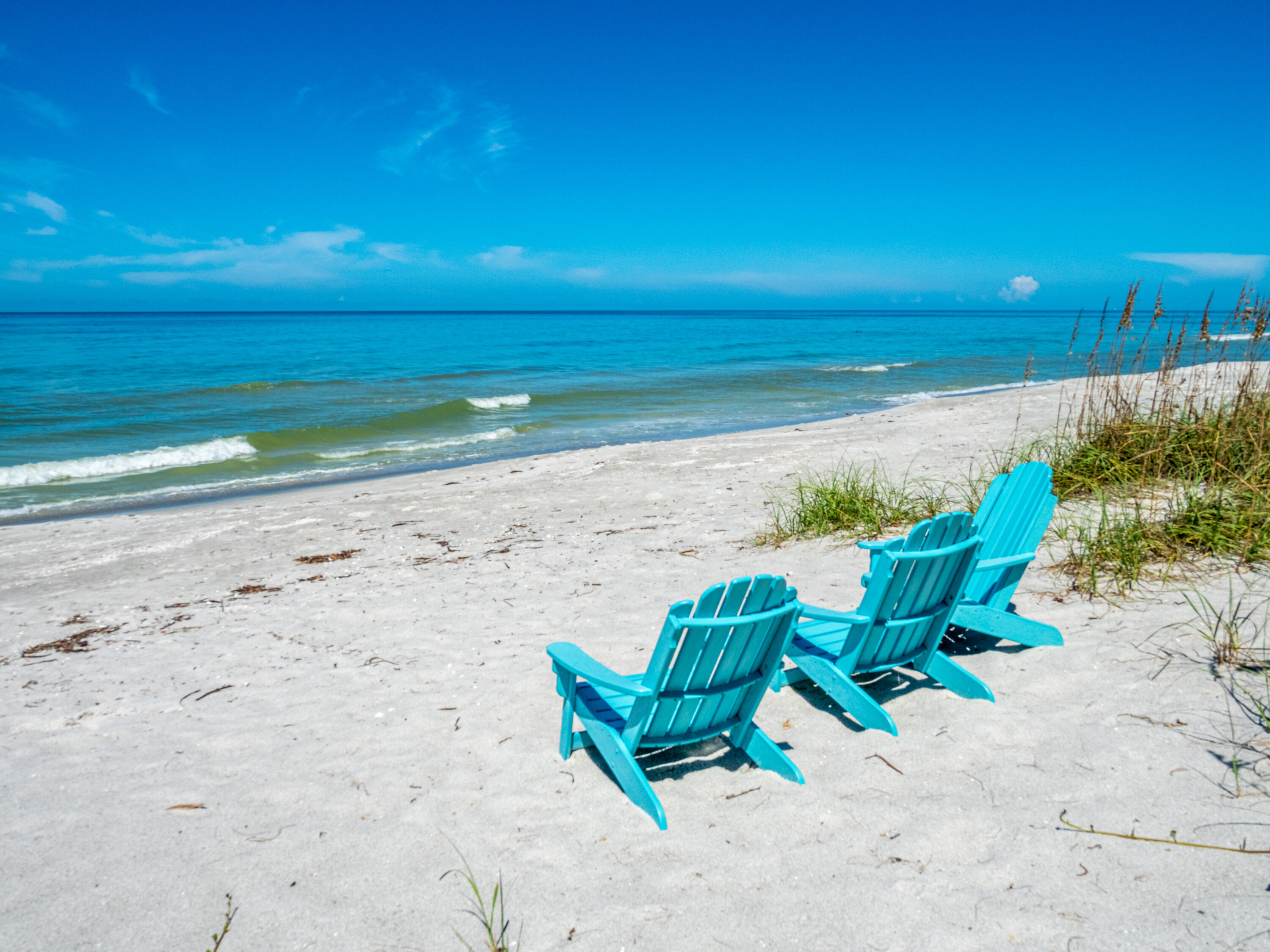 Two blue Adirondack chairs face a tranquil beach with gentle waves under a clear sky.