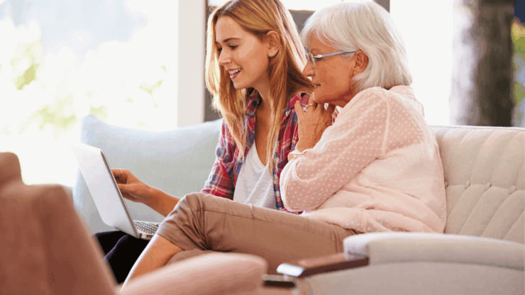 Young woman and elderly woman sit on a sofa, smiling at a laptop.