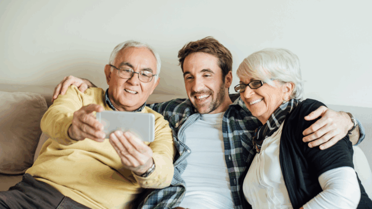 A joyful elderly couple and a young man sit on a couch, smiling as they take a selfie together.