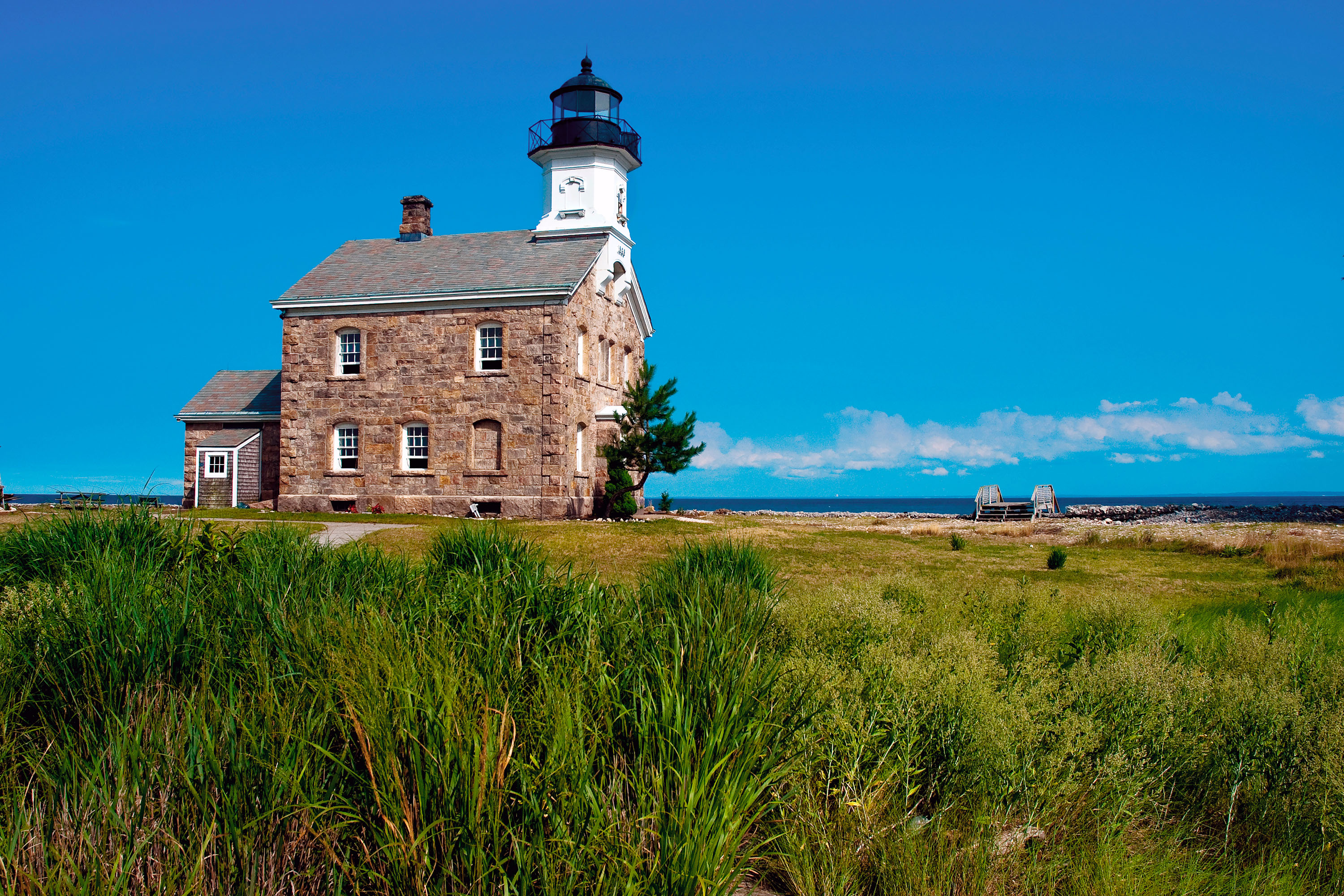 A historic stone lighthouse with a black lantern room, surrounded by grassy land and a view of the sea near Darien, CT.