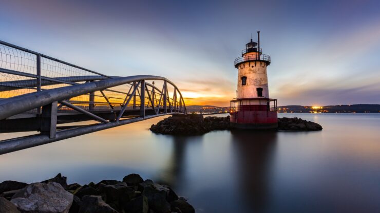 Sleepy Hollow Lighthouse in twilight, connected by a metal bridge, with a colorful sky and calm reflective waters.