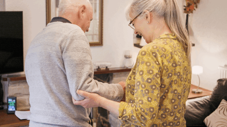 An elderly couple holds hands, smiling warmly in a cozy living room with a TV, lamp, and framed pictures.