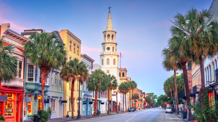 A wide street lined with palm trees, featuring historic buildings in Charleston, South Carolina. The steeple of a church is prominent against a pastel sky during dawn.