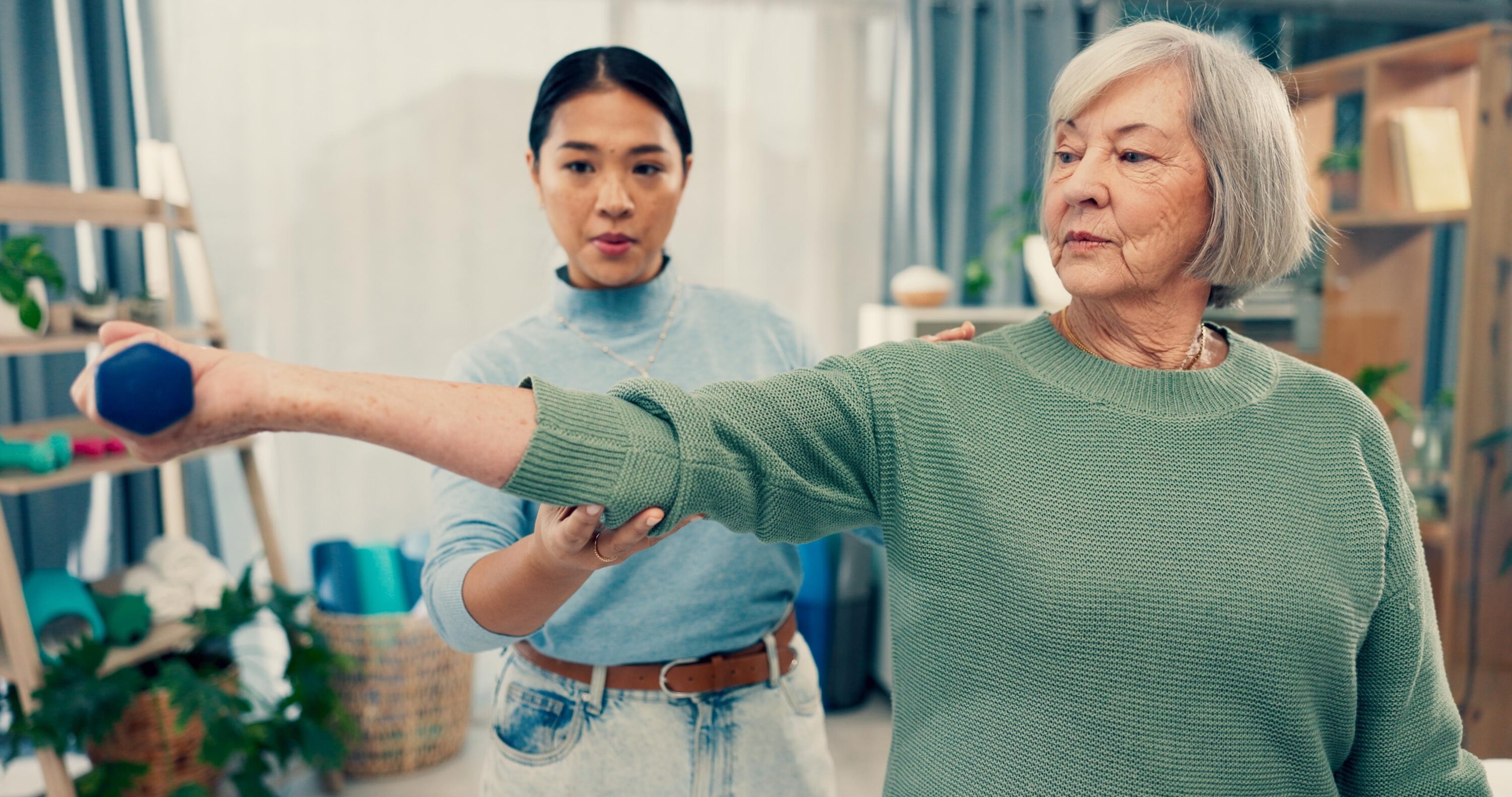 An elderly woman in a green sweater lifts a blue dumbbell, assisted by a younger woman in a light blue shirt.