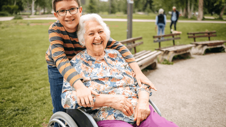 A smiling elderly woman in vibrant floral attire sits in a wheelchair, embraced by a young boy in glasses and stripes. They are outdoors in a park.