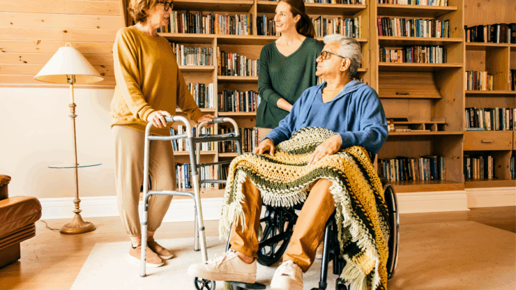 Two women, one with a walker, and an older man in a wheelchair with a knitted blanket, converse happily in a cozy room with bookshelves.