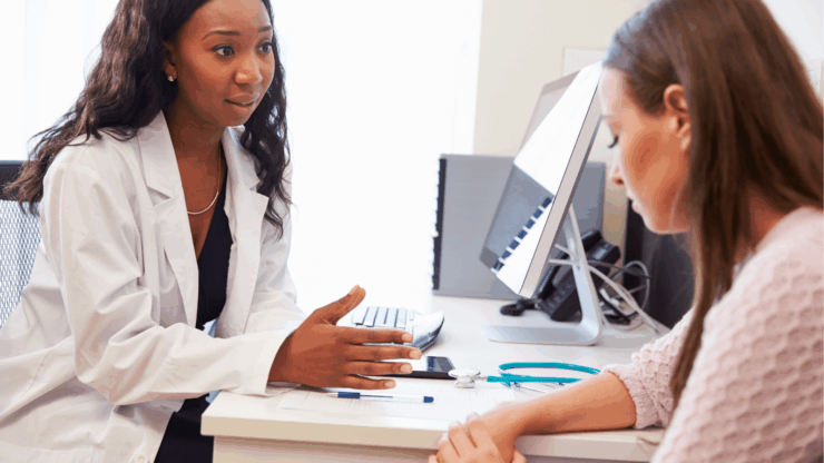 A doctor in a white coat consults attentively with a health care surrogate in an office. A computer and stethoscope are visible, suggesting a medical setting.