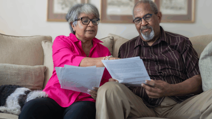 Elderly couple sits on a beige sofa, reviewing paperwork together. The woman wears a bright pink blouse, while the man wears a brown striped shirt.