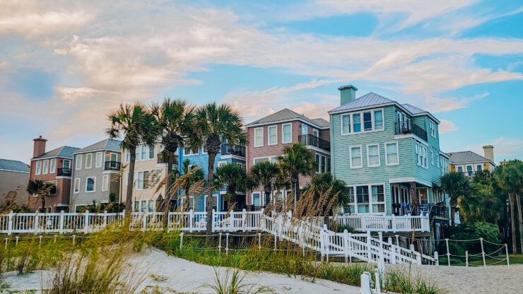 Colorful beach houses with palm trees stand on sandy dunes under a blue sky with wispy clouds.