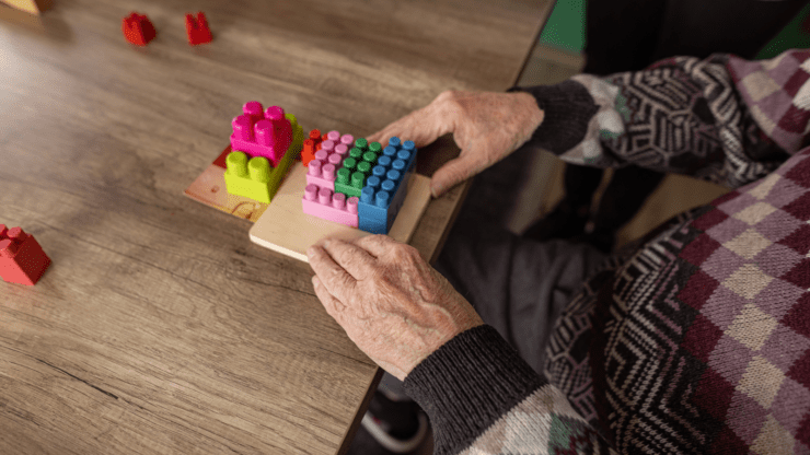 An elderly person wearing a patterned sweater arranges colorful building blocks on a wooden board on a table.