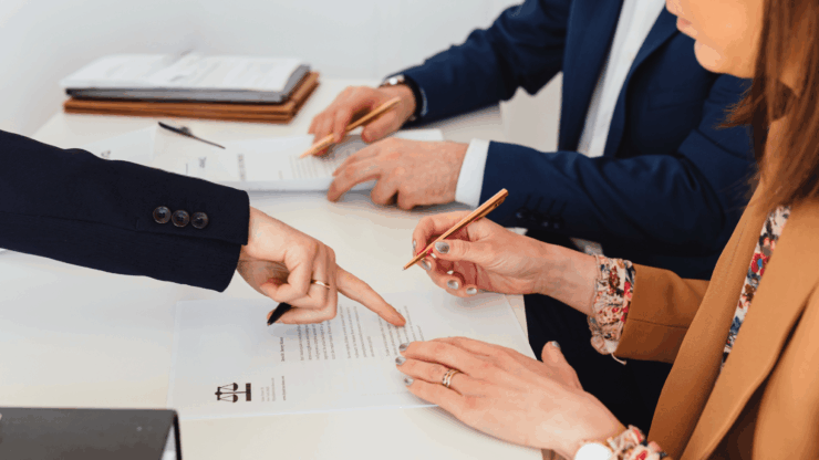 Two professionals reviewing documents at a desk, with a third pointing at a section for clarity.
