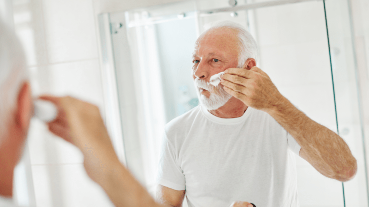 A bearded man in a white T-shirt applies shaving cream to his face, looking into a bathroom mirror.