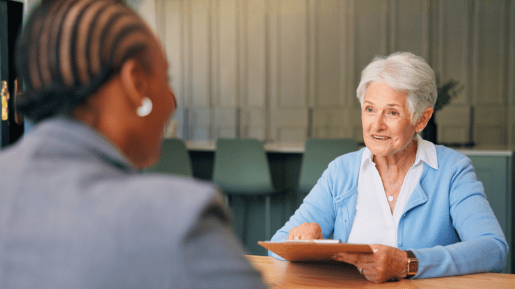 An older woman in a blue cardigan holds a clipboard, conversing with another person in an office setting.