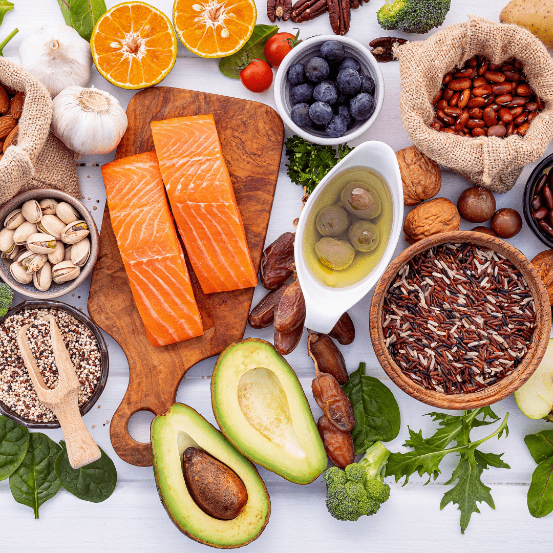 A vibrant display of assorted healthy foods on a white surface, including salmon, avocado, nuts, blueberries, grains, olives, and leafy greens.