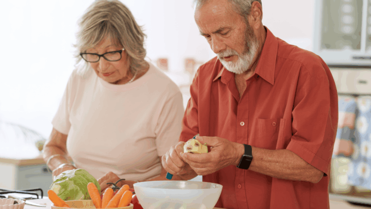 Elderly couple cooking together in a kitchen. Man in red shirt peels an apple over a bowl, while woman in white focuses on vegetables beside him.