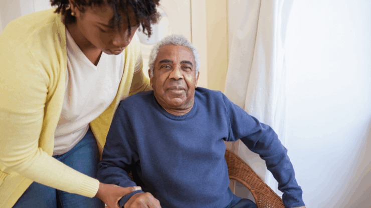 A woman in a yellow sweater assists an elderly man in a blue sweater sitting in a wicker chair.