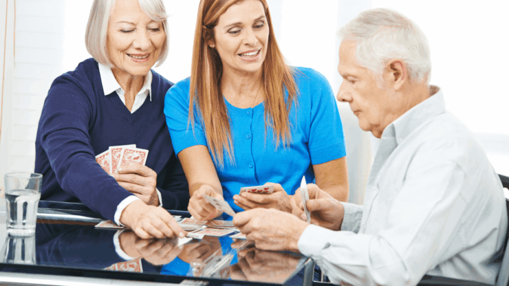 An elderly woman, a younger woman, and an elderly man sit around a glass table, playing a card game together in a bright, indoor setting.