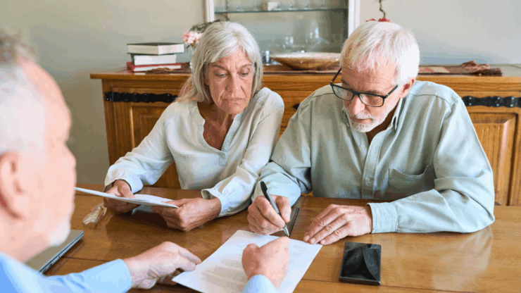 Older couple reviews and signs paperwork at a kitchen table with an adviser, conveying focus and concern during a consultation.