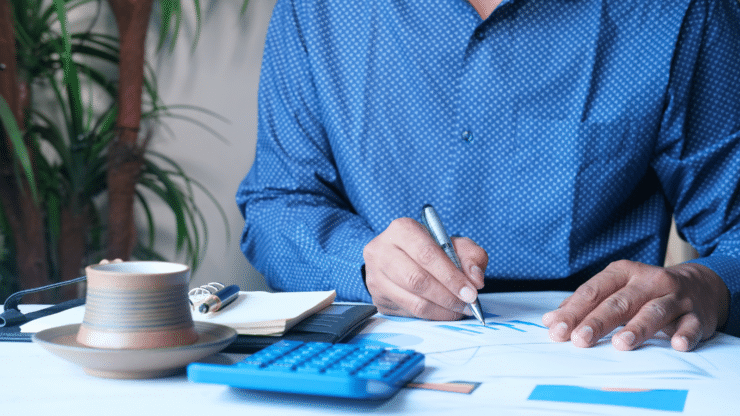 Business professional in a blue shirt signs documents at a desk beside a calculator, coffee, charts, and office plant.