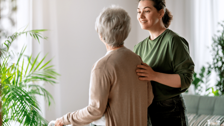 A smiling caregiver gently supports an older woman using a walker in a bright, plant-filled living room.