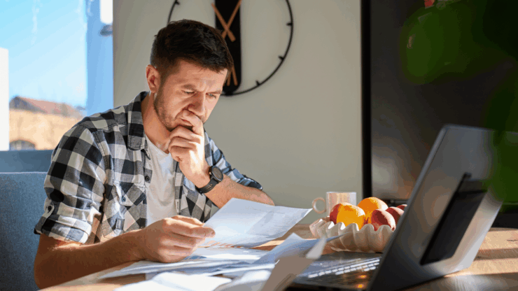 A man studies LTC Insurance paperwork beside an open laptop and fruit bowl, with a large wall clock behind him.