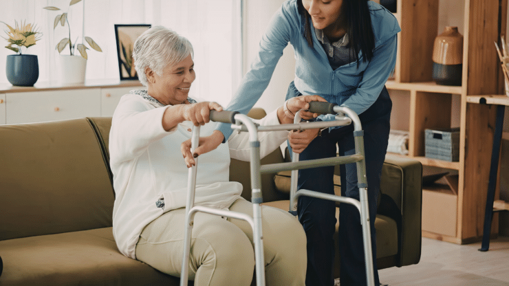 An older woman practices standing with a walker as a caregiver offers steady support.
